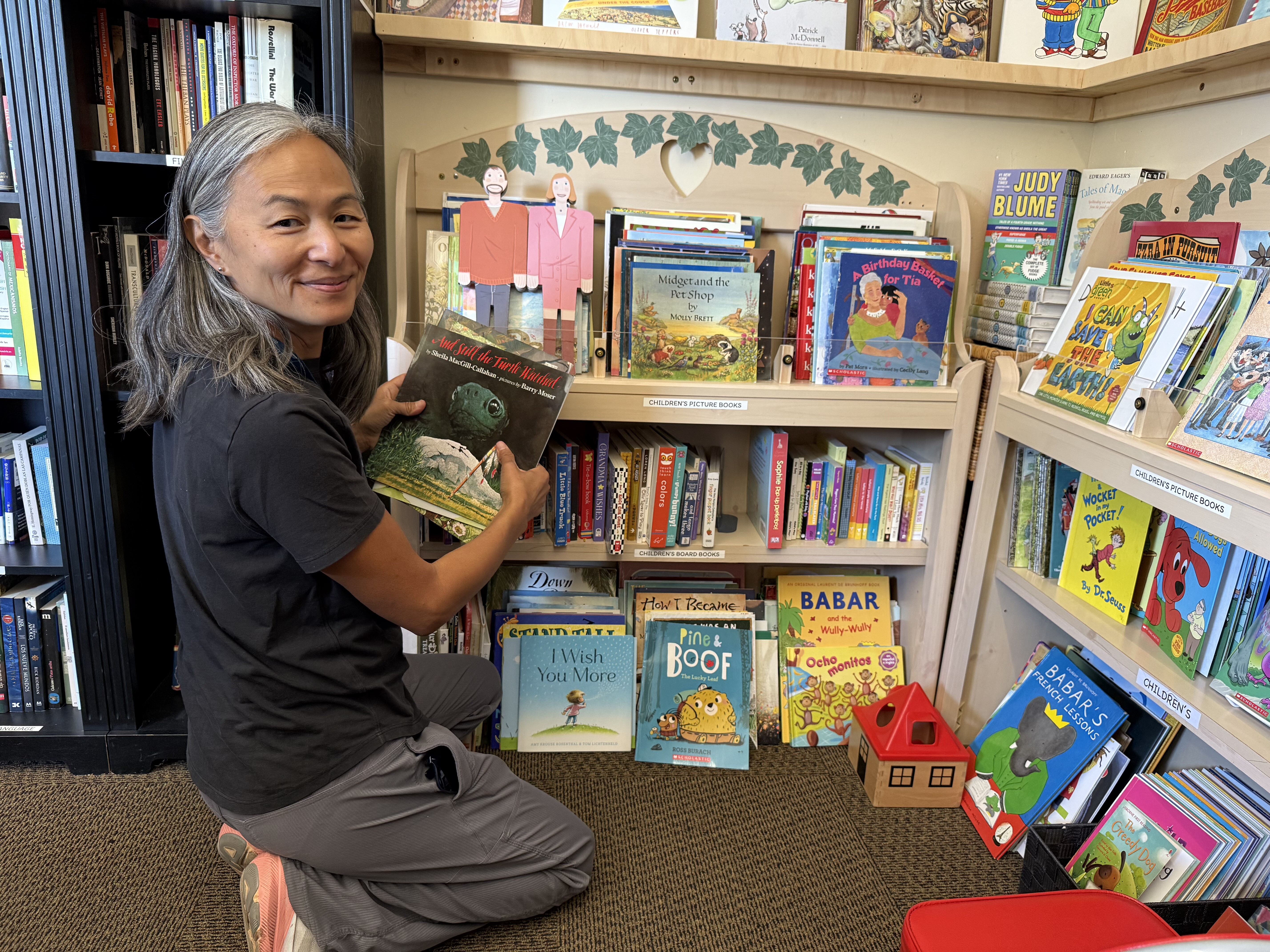 Marcie Soslau Johnson, the new FAL Executive Director, holding a children's book and kneeling next to a bookshelf in the Books for Friends bookshop.
